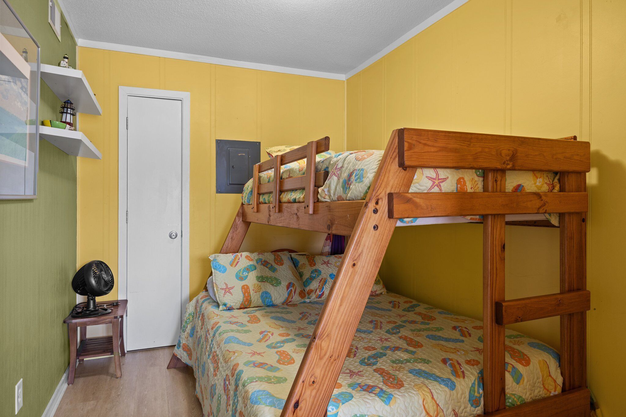 A bunk bed with a wooden frame and colorful bedding, a small table with a fan, and a door with a light switch.