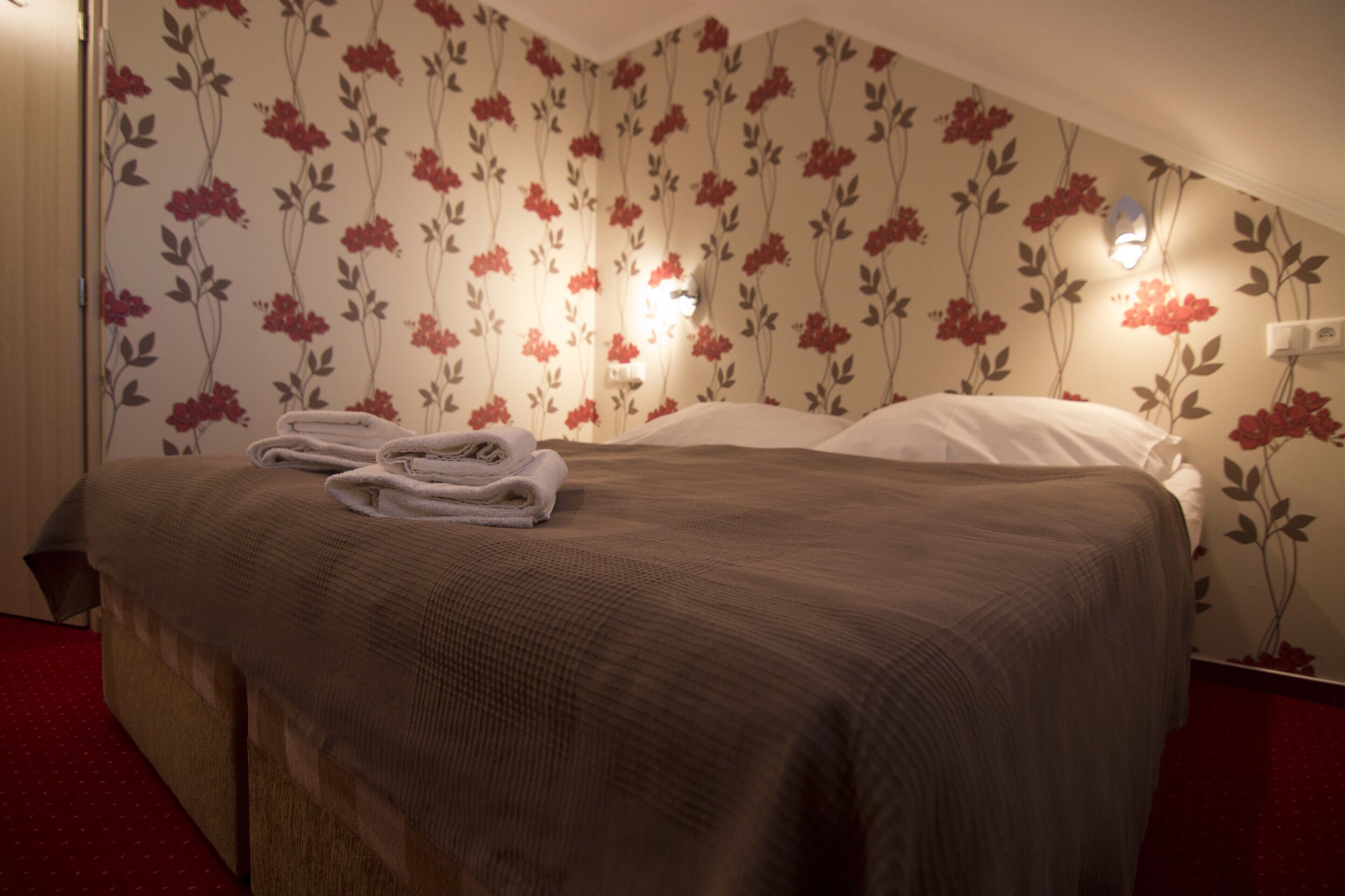 Een net opgemaakt bed met een bruine deken en witte kussens, een opgevouwen handdoek op het bed, en een bloemenpatroon op het behang.