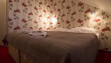 Een net opgemaakt bed met een bruine deken en witte kussens, een opgevouwen handdoek op het bed, en een bloemenpatroon op het behang.
