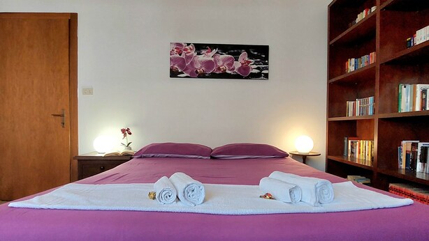 Une chambre d’hôtel avec un grand lit, une literie violette, une bibliothèque remplie de livres et un tableau représentant des fleurs accroché au mur.