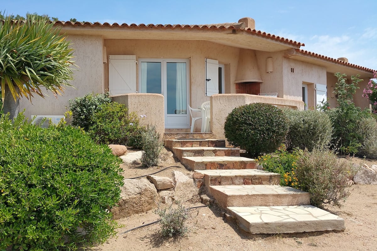 Une maison à l’extérieur beige, avec un toit en tuiles et une porte d’entrée dotée d’un panneau de verre. Des marches mènent à la porte, qui est entourée de différentes plantes et arbustes.