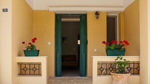 Fachada de edificio color beige, puerta verde y macetas con flores rojas a ambos lados.