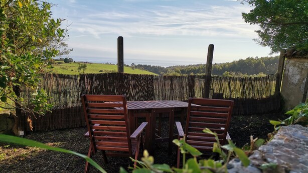 Ein Holztisch und zwei Stühle in einem Garten mit Blick auf ein Feld und Bäume in der Ferne.