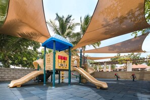 A playground with slides and climbing structures, surrounded by palm trees and shaded by large canopies.