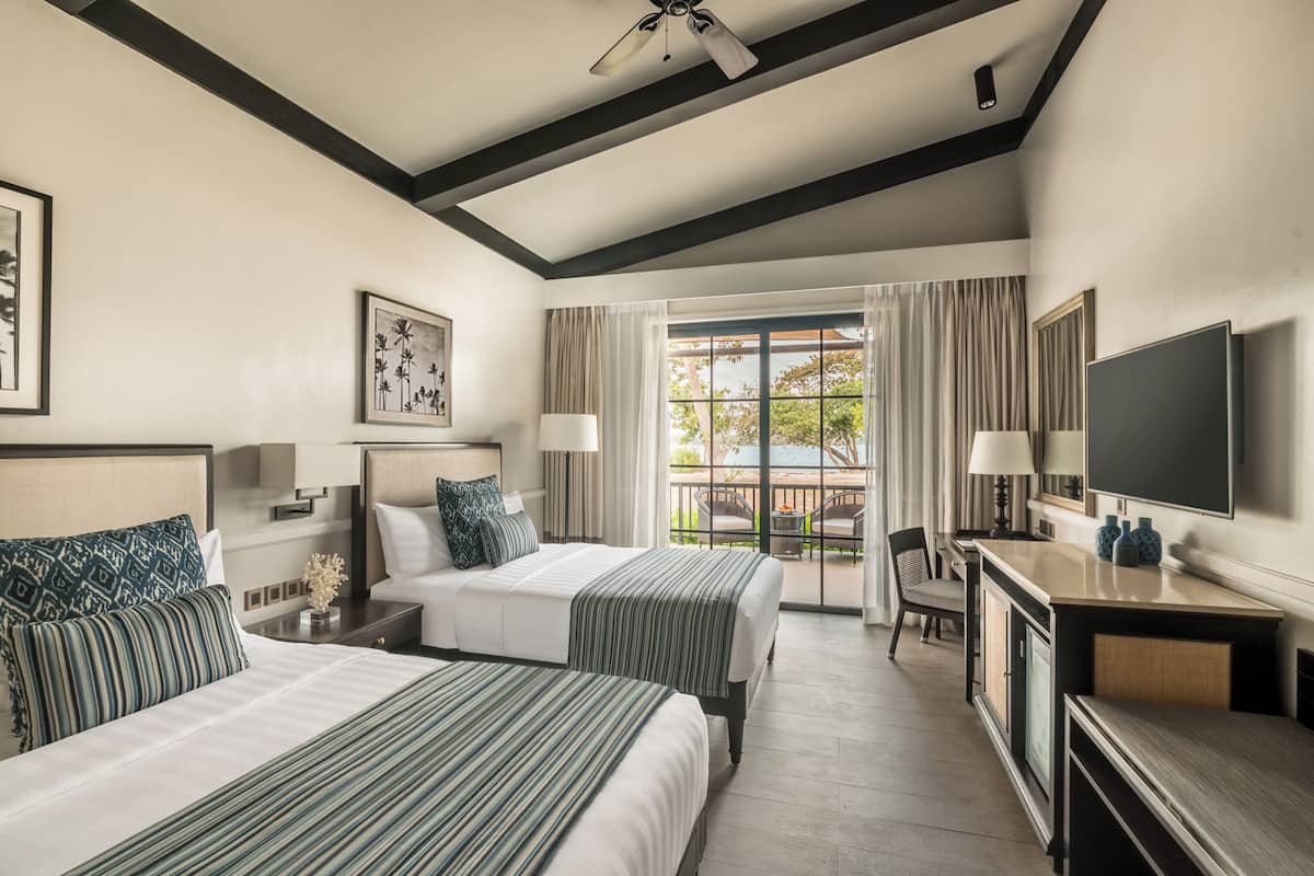 A hotel room with two beds, a desk, a TV, and a balcony with curtains.