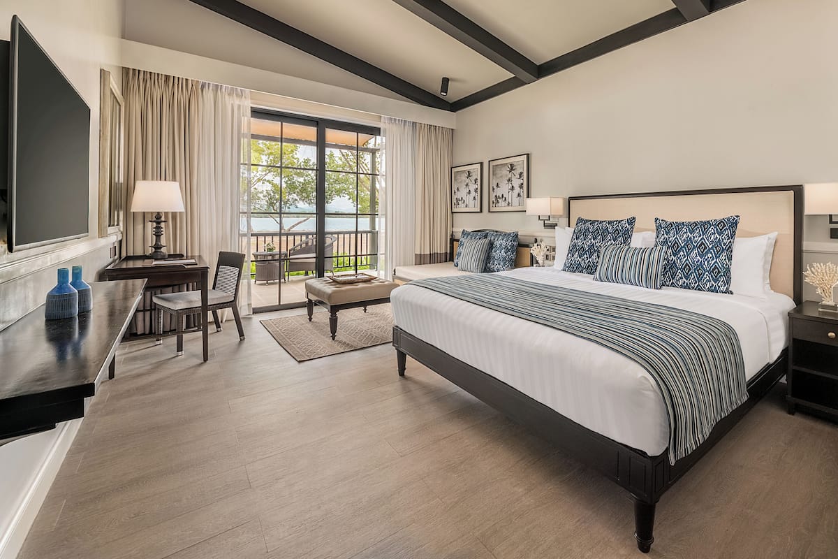 A hotel room with a large bed, a desk, a chair, a balcony with curtains, and a ceiling fan.