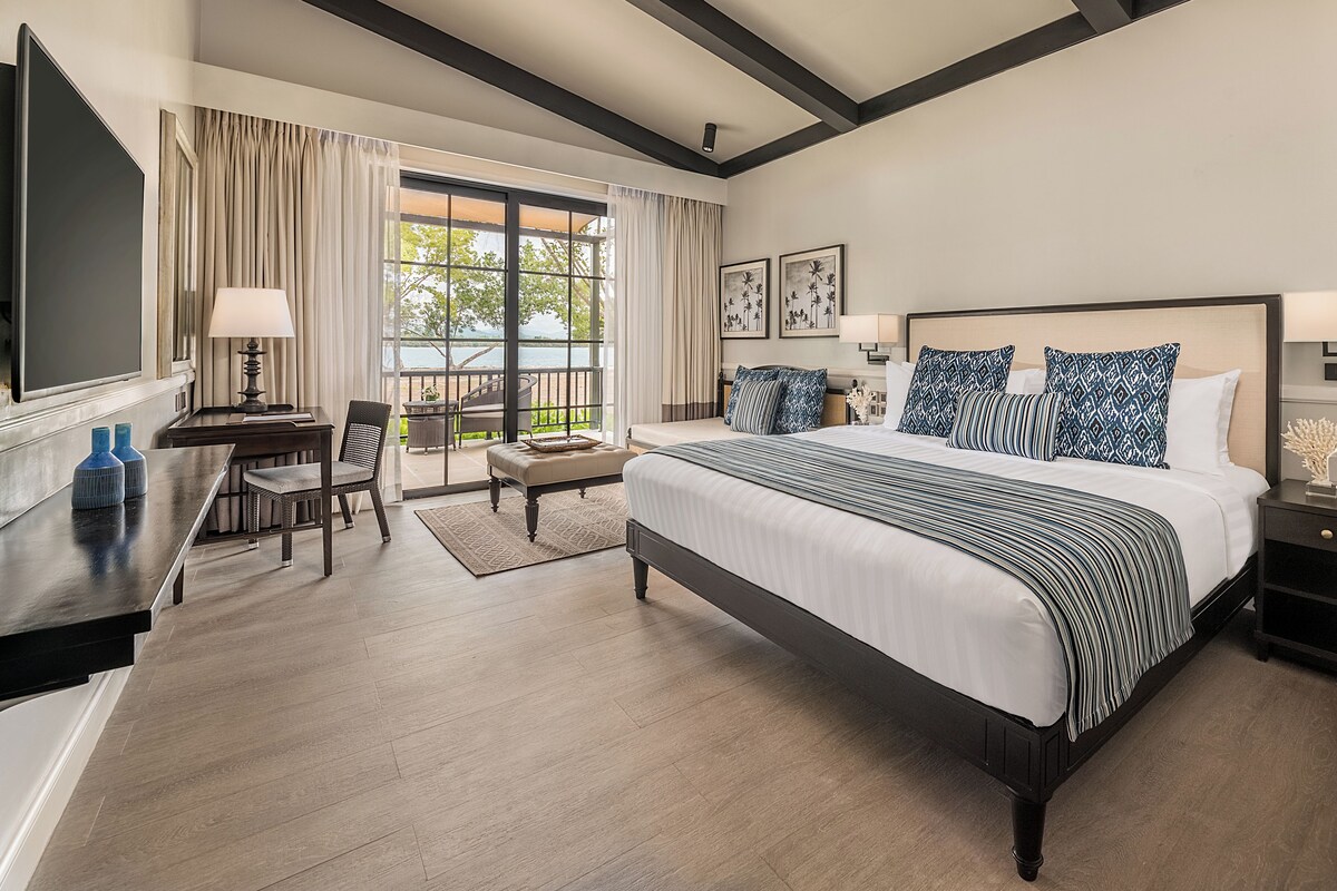 A hotel room with a large bed, a desk with a chair, a balcony with curtains, and a ceiling fan.