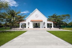A white church with a triangular roof and a central circular window, surrounded by a paved walkway and palm trees.