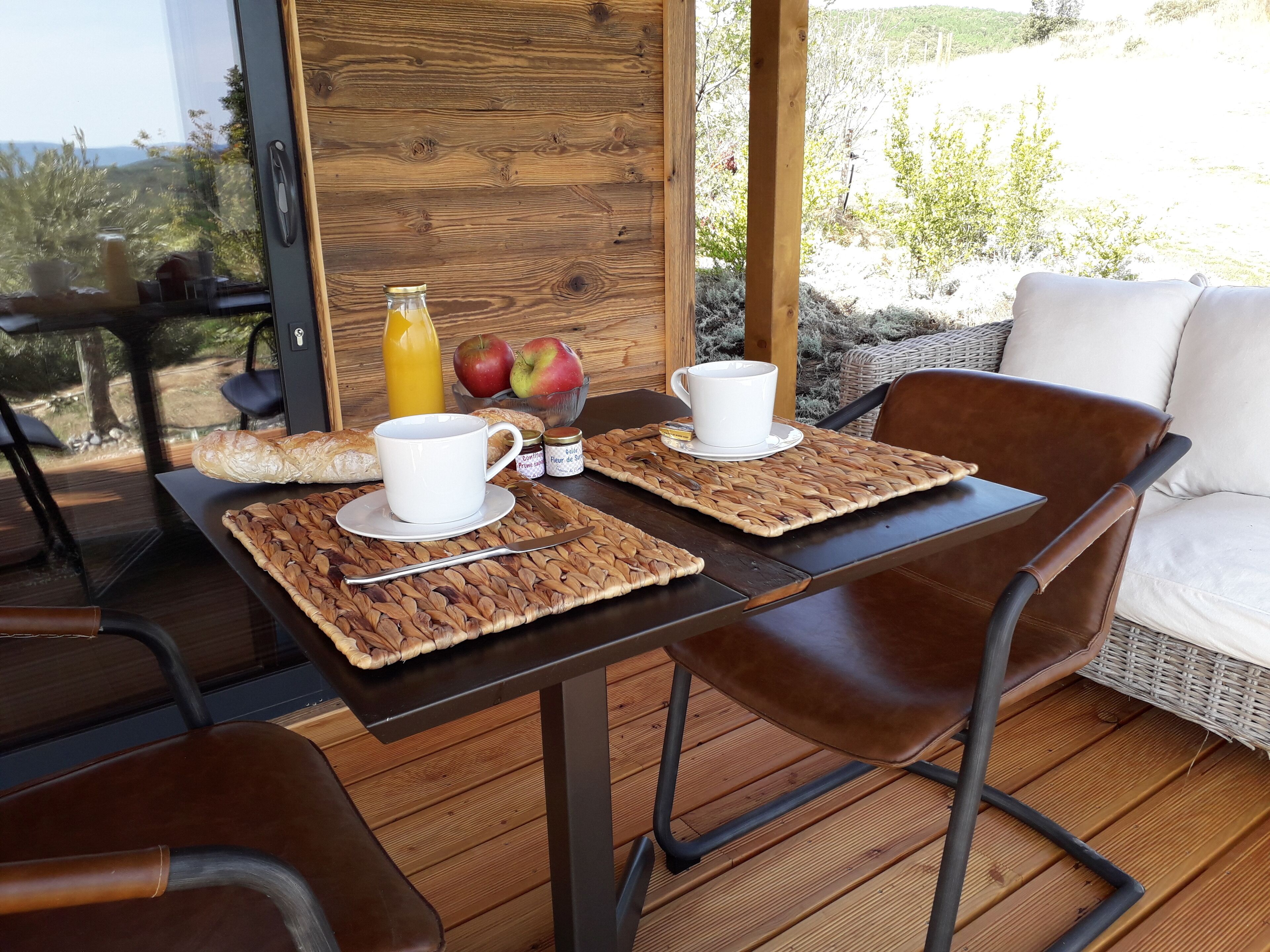 A wooden deck with two tables, each with a white cup and saucer, a baguette, and a bottle of orange juice.