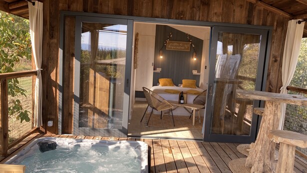 Una cabaña de madera con jacuzzi, un comedor con mesa y sillas, y un dormitorio con cama y cortinas.