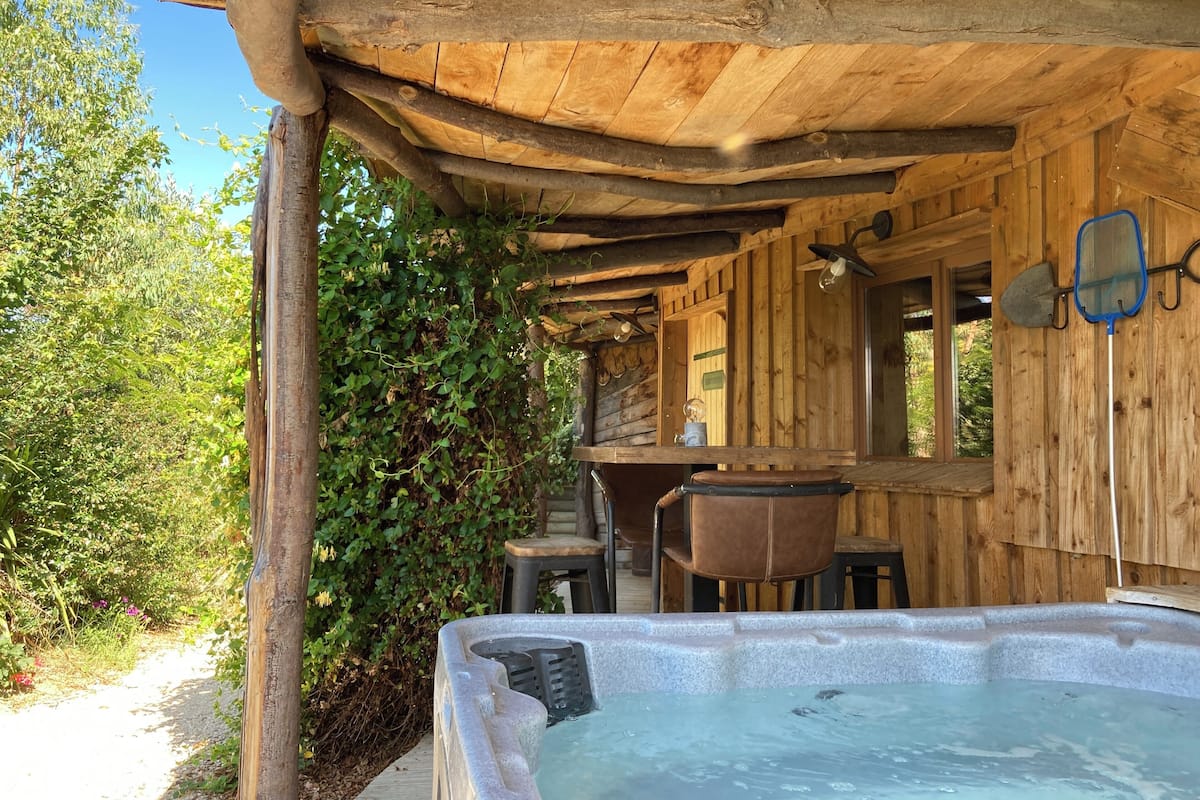 A wooden cabin with a hot tub, wooden walls, and a window overlooking greenery.