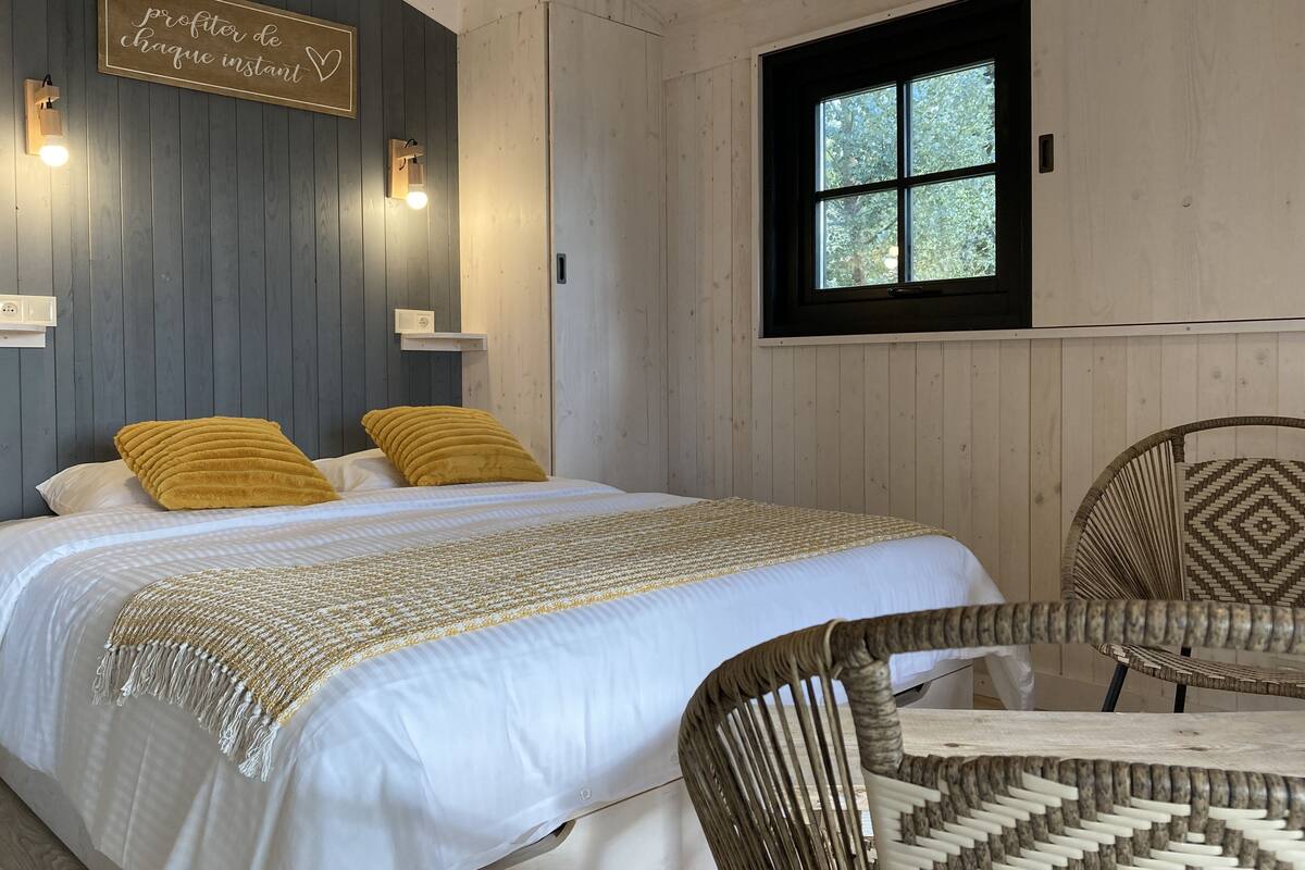 A bedroom with a bed, two yellow pillows, a bedside table, a wall-mounted light, and a window with a view of trees.