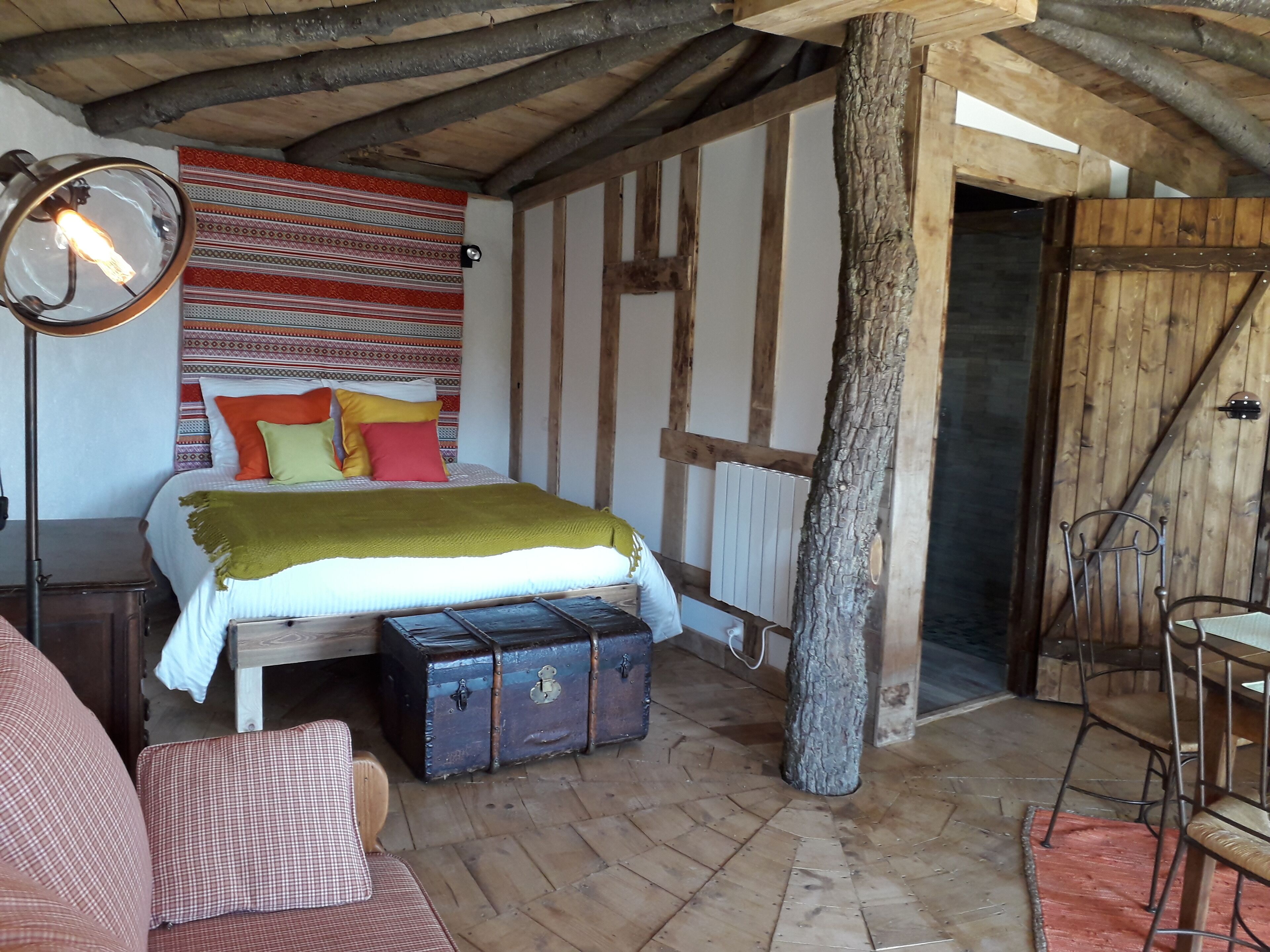 A rustic bedroom with a bed, a chest of drawers, a wooden floor, and a large tree trunk.