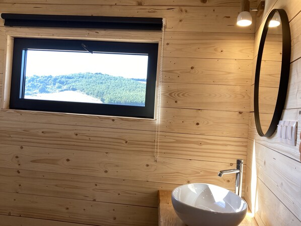 A wooden cabin bathroom with a large window, a round mirror, and a vessel sink. - Cabanes Trésors de Campagne (Villarzel-du-Razes)