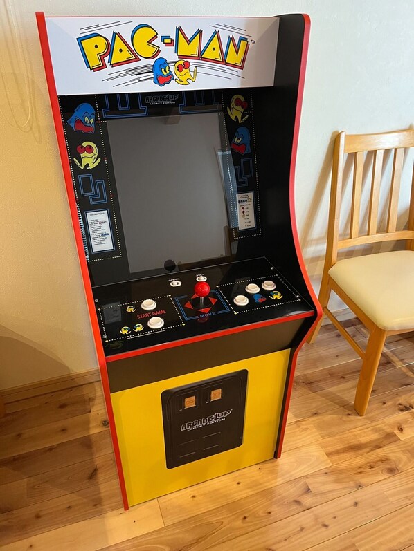 Une borne d’arcade Pac-Man dans une pièce dont le sol est recouvert de parquet et qui contient une chaise en bois.