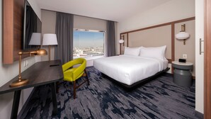 A hotel room with a bed, a desk with a lamp, a chair, a bedside table with a phone, and a large window with a city view. - Fairfield by Marriott® Inn & Suites San Luis Potosi (San Luis Potosi)