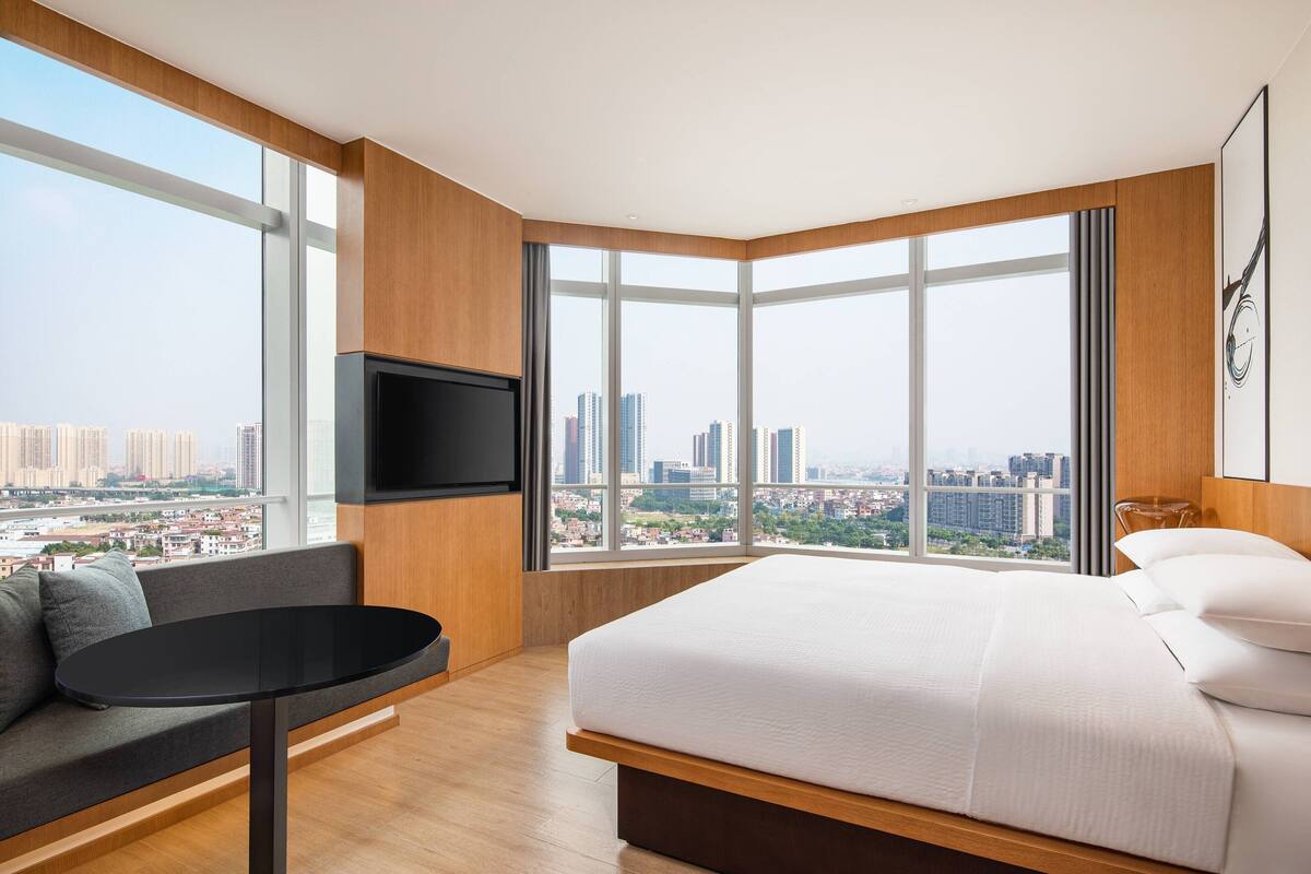 Ein modernes Hotelzimmer mit einem großen Bett, einer Sitzecke, einem an der Wand befestigten Fernseher und Blick auf die Stadt durch große Fenster.