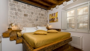 Ein rustikales Holzgestellbett vor einer Steinwand, ein Fenster mit Fensterläden und ein Bild an der Wand.