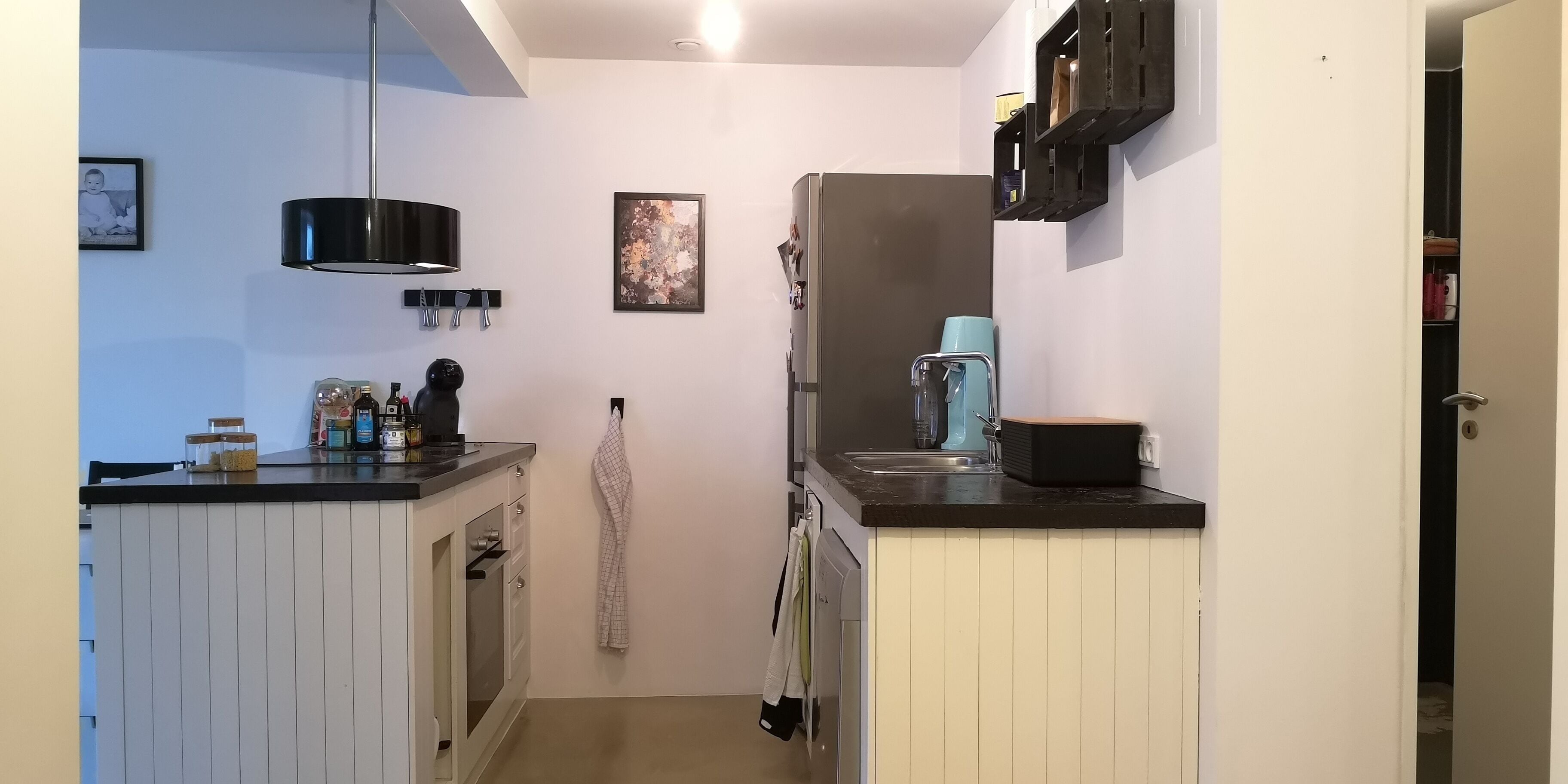 A narrow kitchen with white cabinets, a black countertop, a hanging light, and a refrigerator.