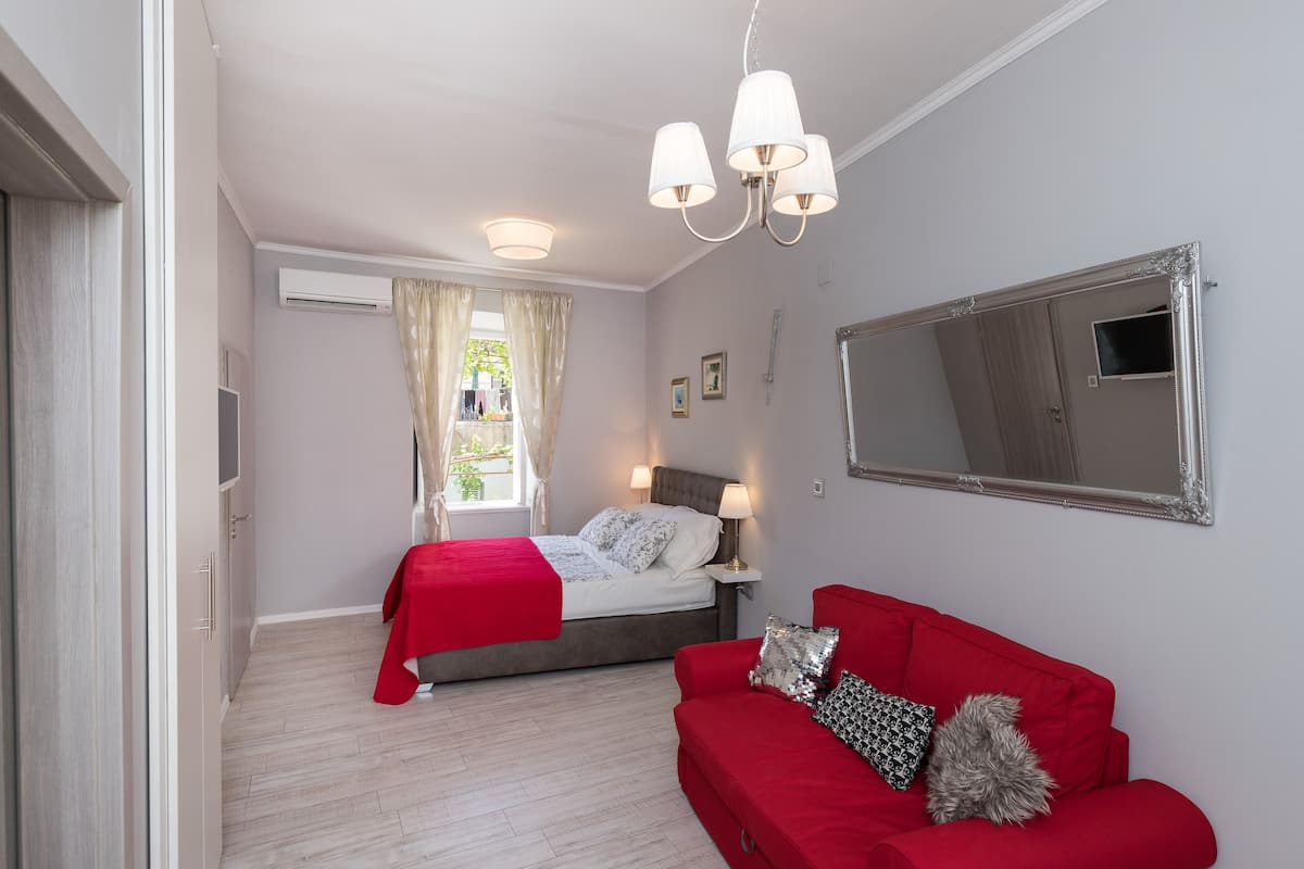 Quarto com cama, sofá vermelho, espelho grande e janela com cortinas.