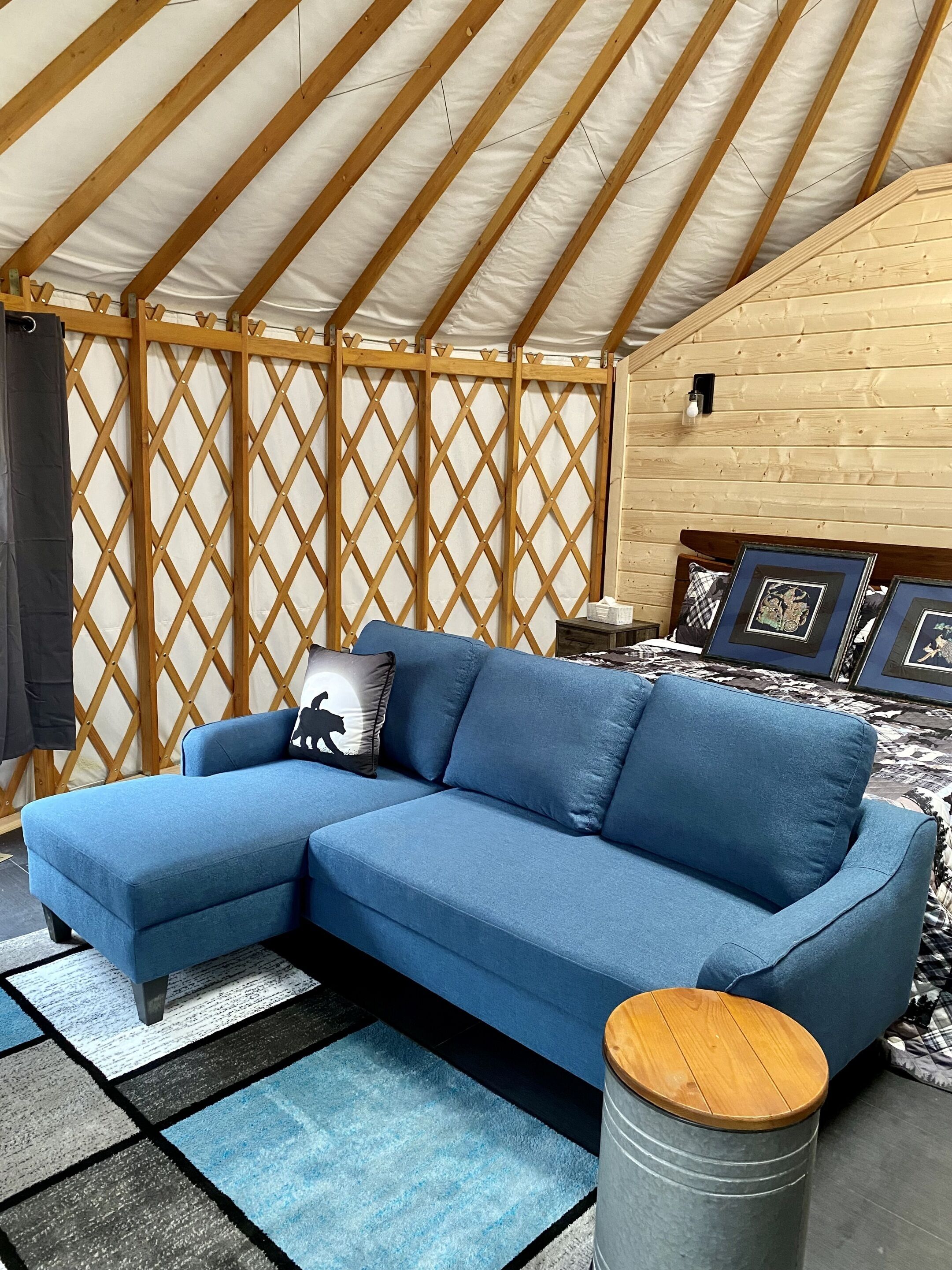 A blue sectional sofa with a black silhouette pillow, a wooden stool, and a patterned rug in a tent-like room with wooden beams and lattice walls.