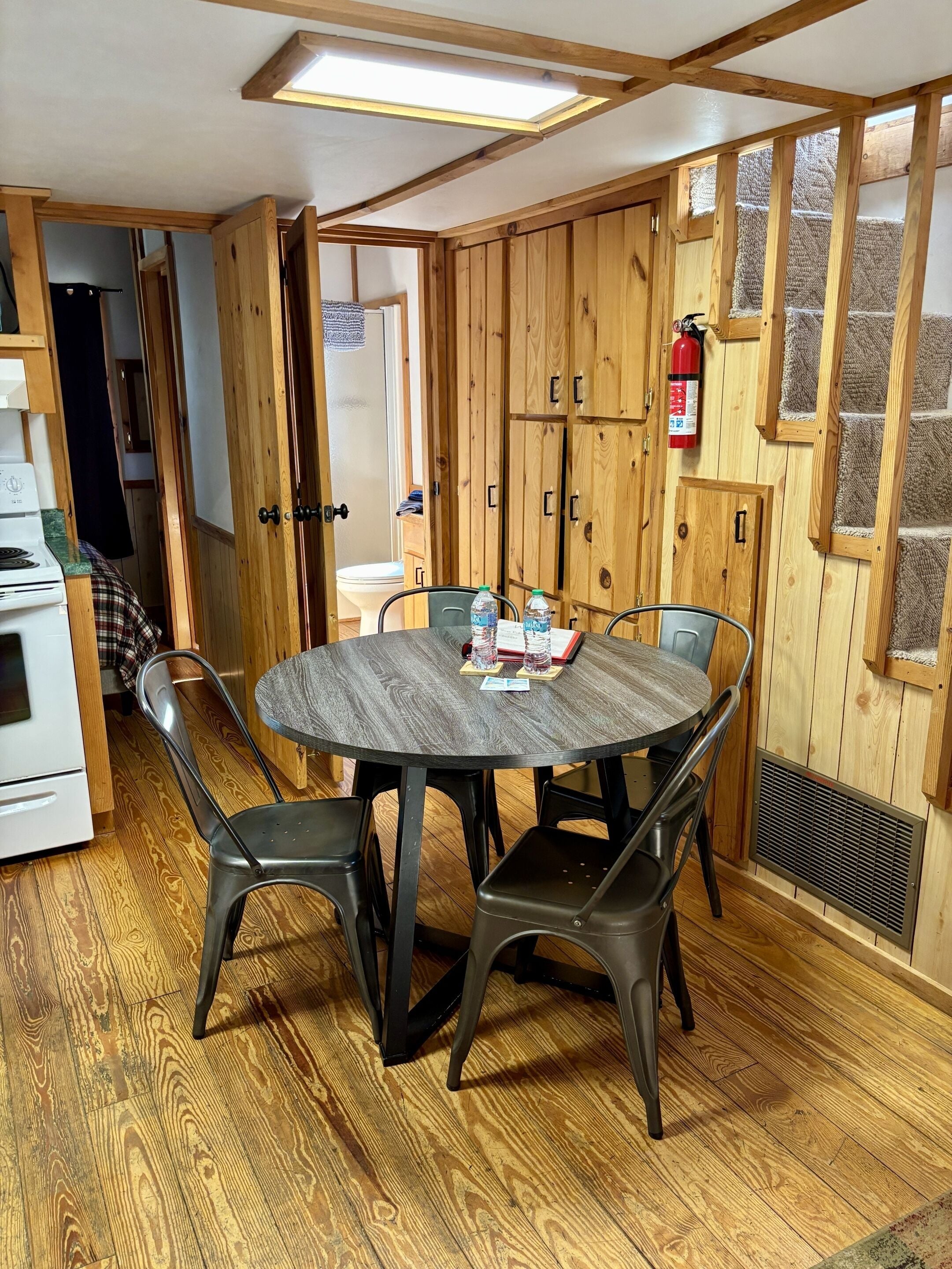 Una habitación con una mesa redonda, cuatro sillas, paredes de madera, un extintor y una rejilla de ventilación.
