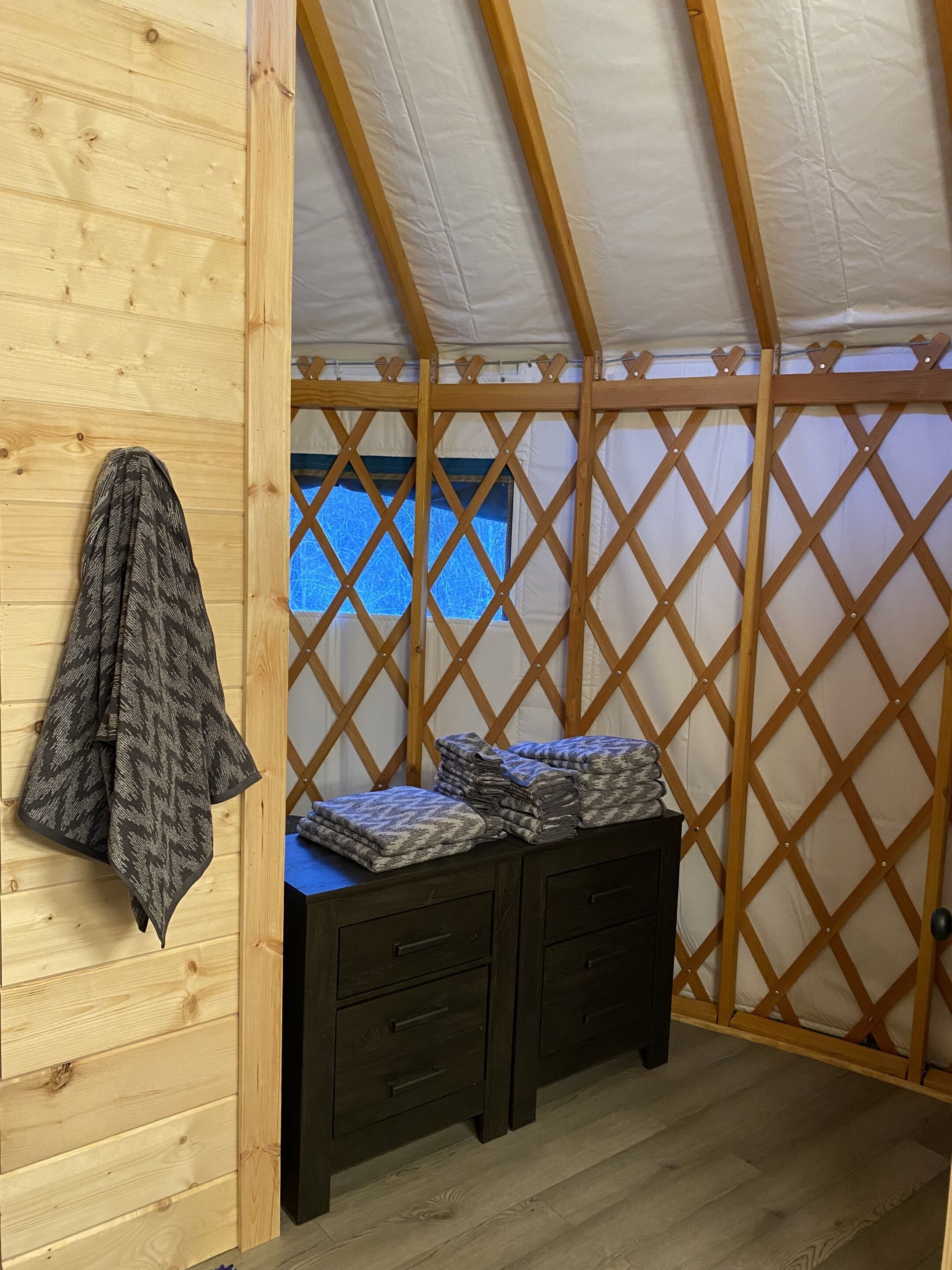 Una stanza con soffitto spiovente, un divisorio in legno intagliato, un letto con materasso blu, una cassettiera in legno scuro e una coperta decorata appesa al muro.