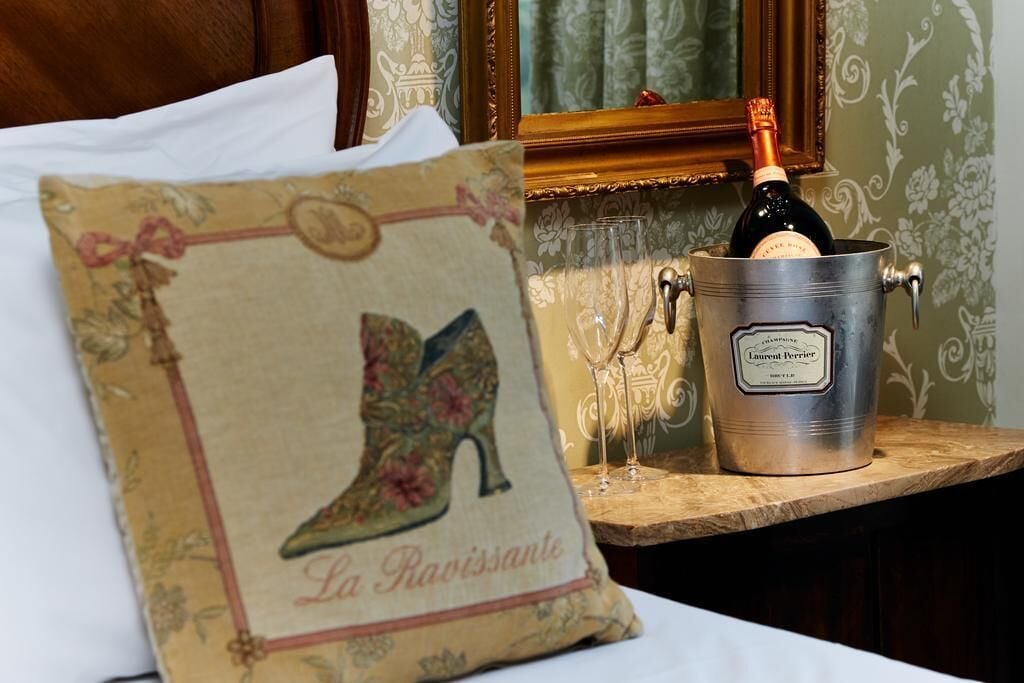 Un almohilar con diseño de bota y el texto "La Ravissante" sobre una cama. Un balde de metal con champán y copas está sobre una mesa de madera.