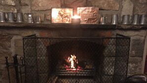 Une cheminée en pierre avec un foyer en métal, un feu allumé et une étagère au-dessus où sont posées plusieurs tasses en métal.
