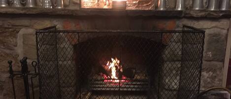 A stone fireplace with a metal grate, a lit fire, and a shelf above holding several metal cups.