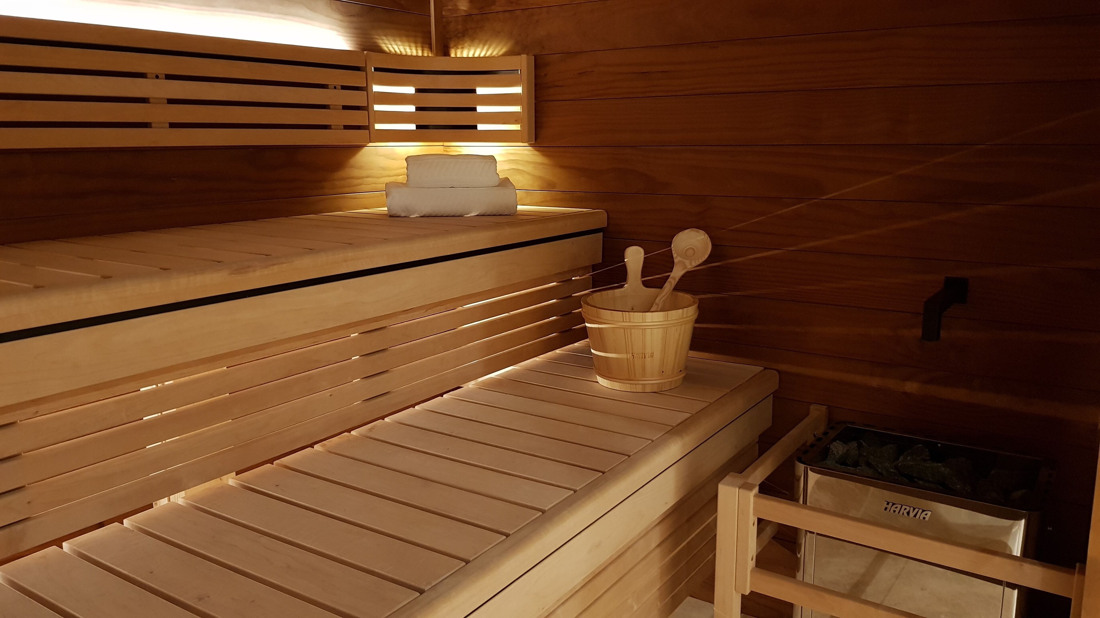 Un sauna en bois avec des bancs, un seau en bois avec une louche et un appareil de chauffage pour sauna de la marque Harvia.