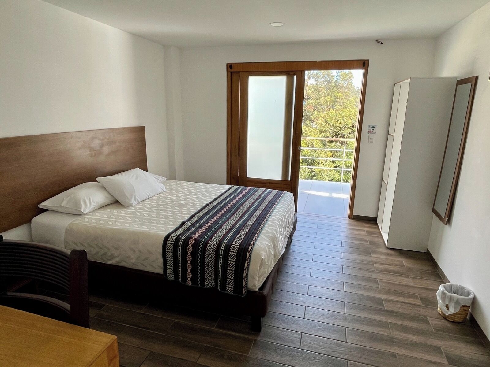 Un dormitorio con cama, cabecera de madera, manta estampada, piso de madera y vista a la vegetación a través de una puerta corrediza.