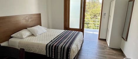 Un dormitorio con cama, cabecera de madera, manta estampada, piso de madera y vista a la vegetación a través de una puerta corrediza.