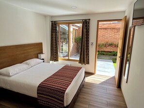 A bedroom with a large bed, a wooden headboard, a bedside table, and a window with curtains. - Hostal El Geranio (Otavalo)