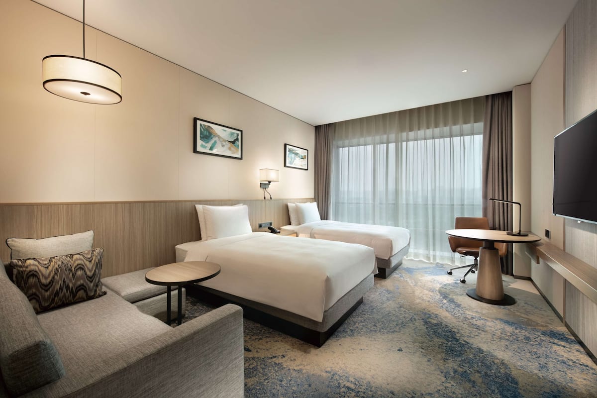 A hotel room with two beds, a sofa, a small table, a TV, and a large window with curtains.