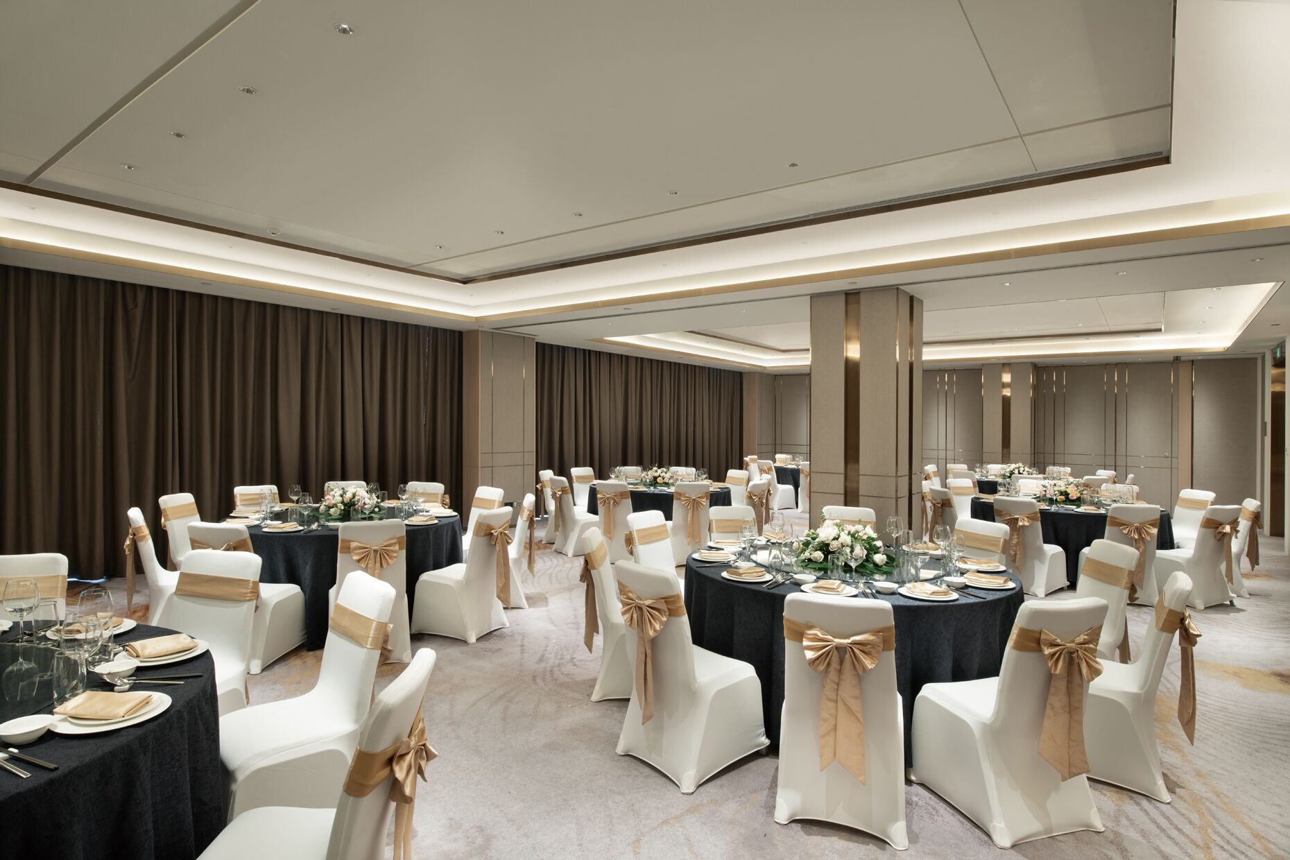 A banquet hall with round tables, white chairs with gold sashes, and dark tablecloths.
