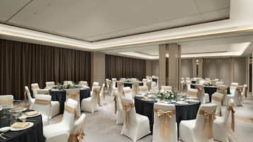 A banquet hall with round tables, white chairs with gold sashes, and dark tablecloths.