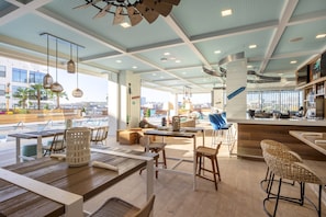 Un restaurante moderno con mesas de madera y sillas de mimbre, una zona de bar y grandes ventanales con vistas a la ciudad.