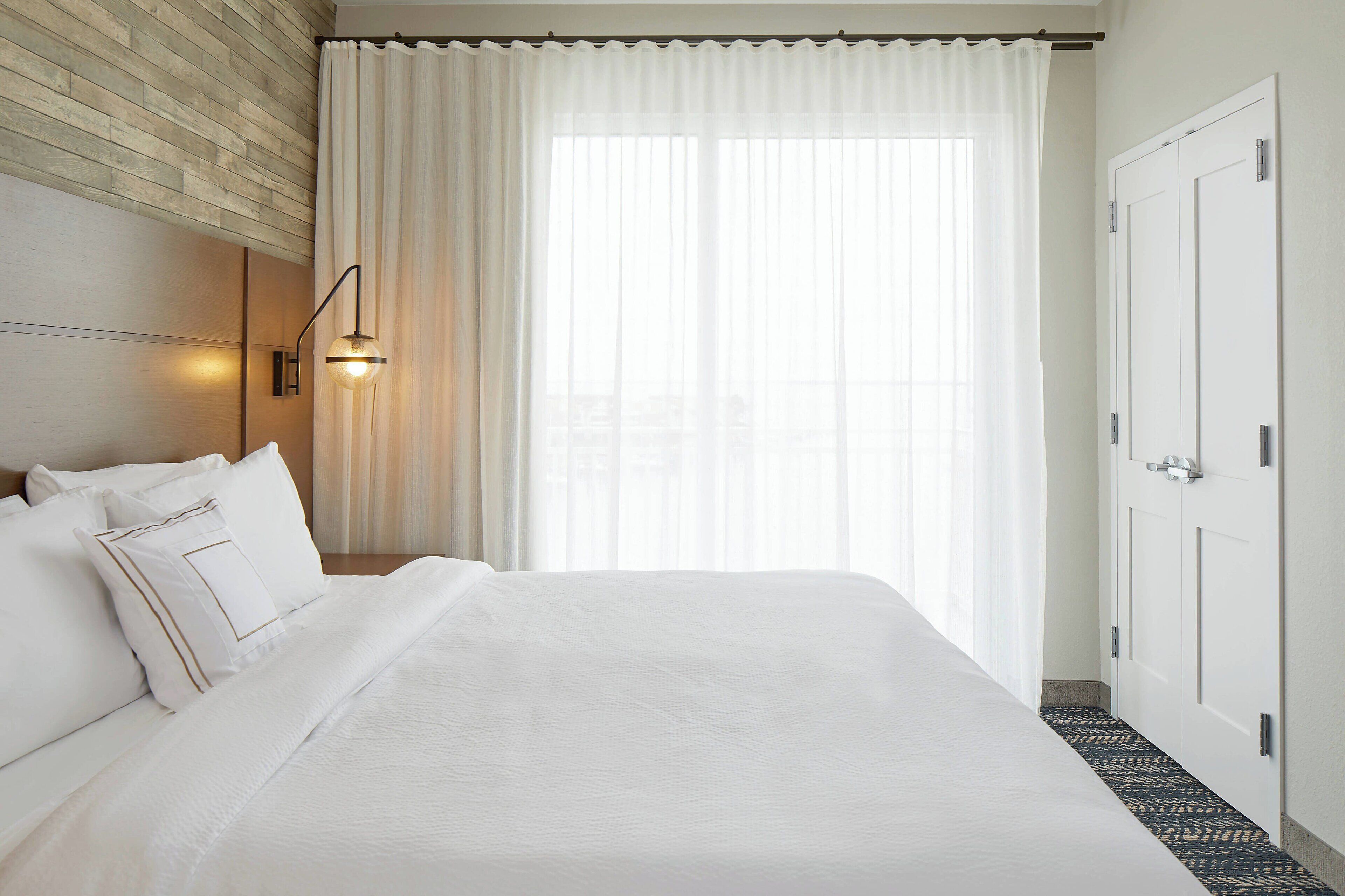 Hotellihuone, jossa on suuri sänky, pääty, seinään kiinnitetty valaisin ja ovi.