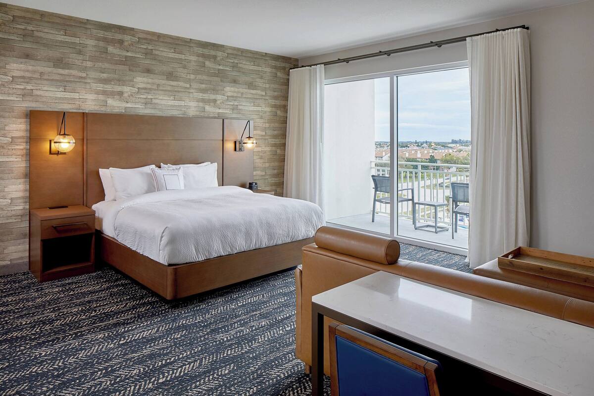 A hotel room with a large bed, a nightstand, a desk, and a view of the city through a large window.