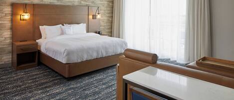 Une chambre d’hôtel avec un grand lit, une tête de lit en bois, une table de chevet, un bureau et une fenêtre avec des rideaux légers.