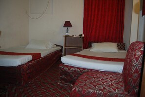 A hotel room with two beds, a red patterned chair, a small table with a lamp, and a red curtain.