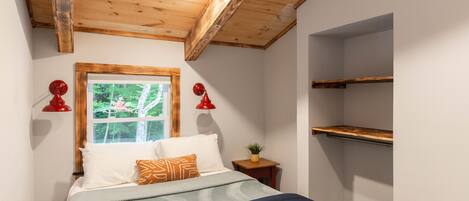 A bedroom with a wooden ceiling, a bed with a blue bedspread, two red wall lamps, a window with a view of trees, and a small wooden nightstand with a plant.