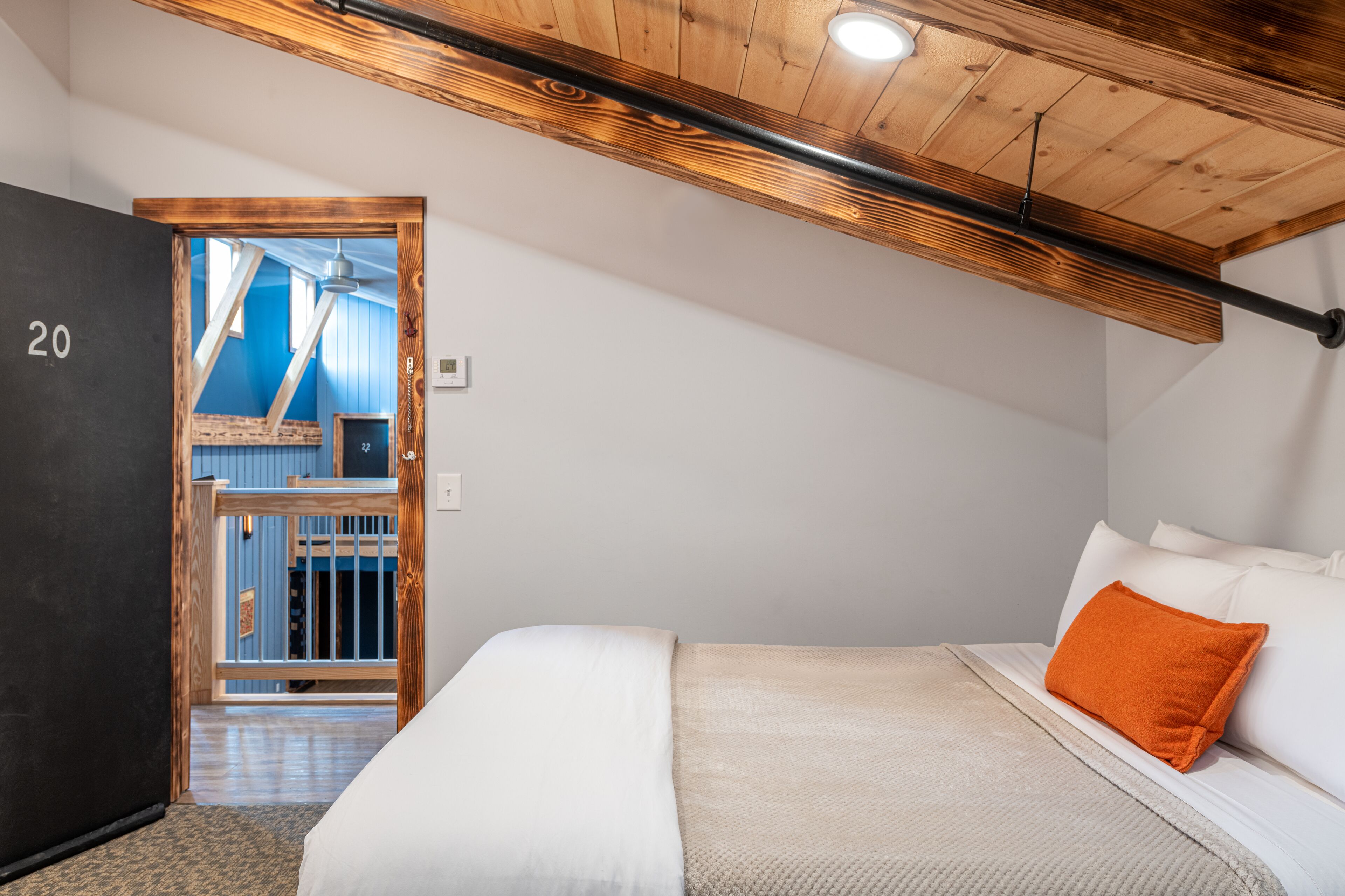 Una camera da letto con un letto, un soffitto in legno, una porta con un numero e vista su una stanza blu attraverso una porta vetrata.