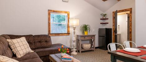 A cozy living room with a brown sofa, a wooden coffee table with books and a vase of flowers, a lamp, and a dining area with a table set for two.