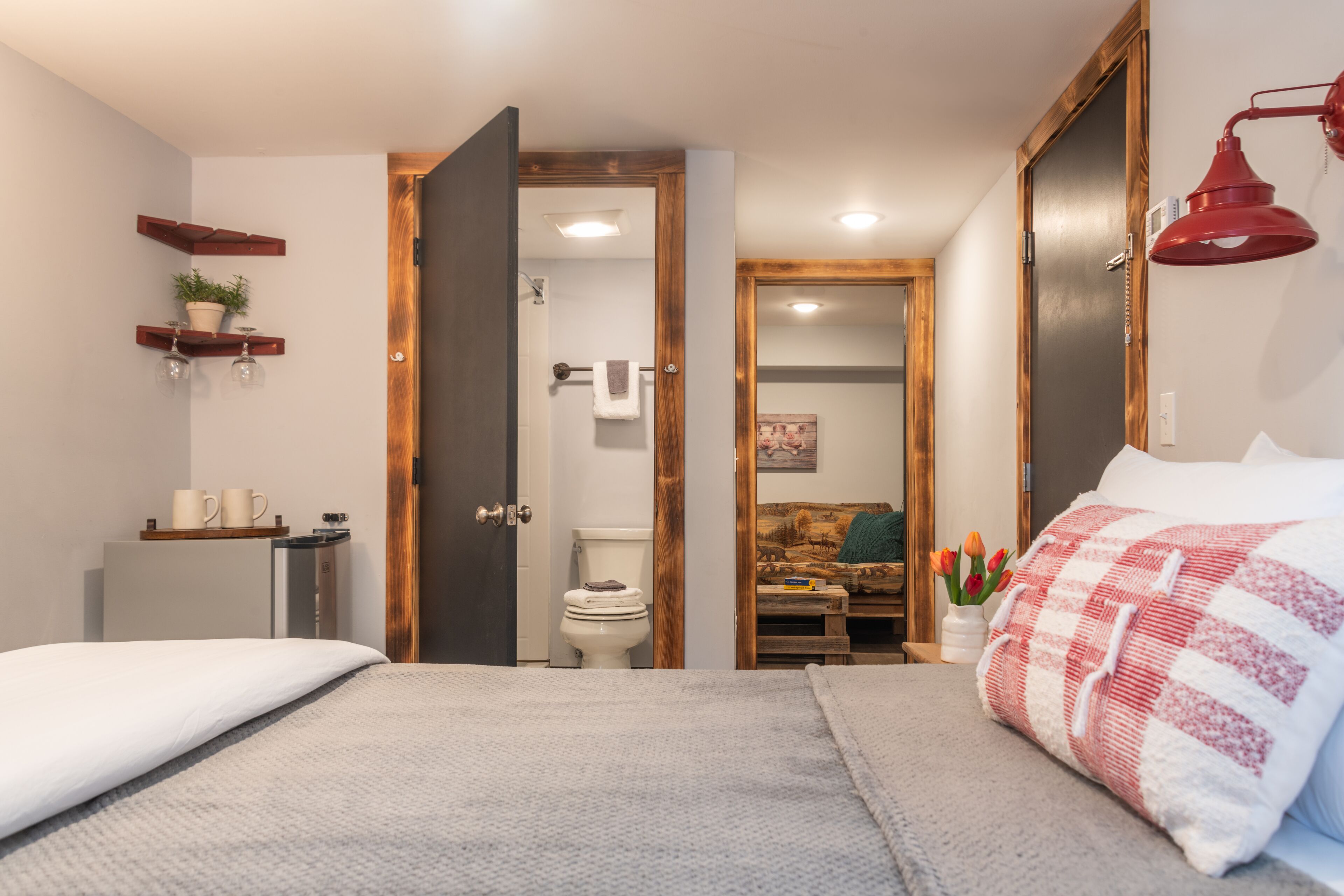 A bedroom with a bed, a bedside table, a nightstand, a red lamp, and a wooden door leading to a bathroom.