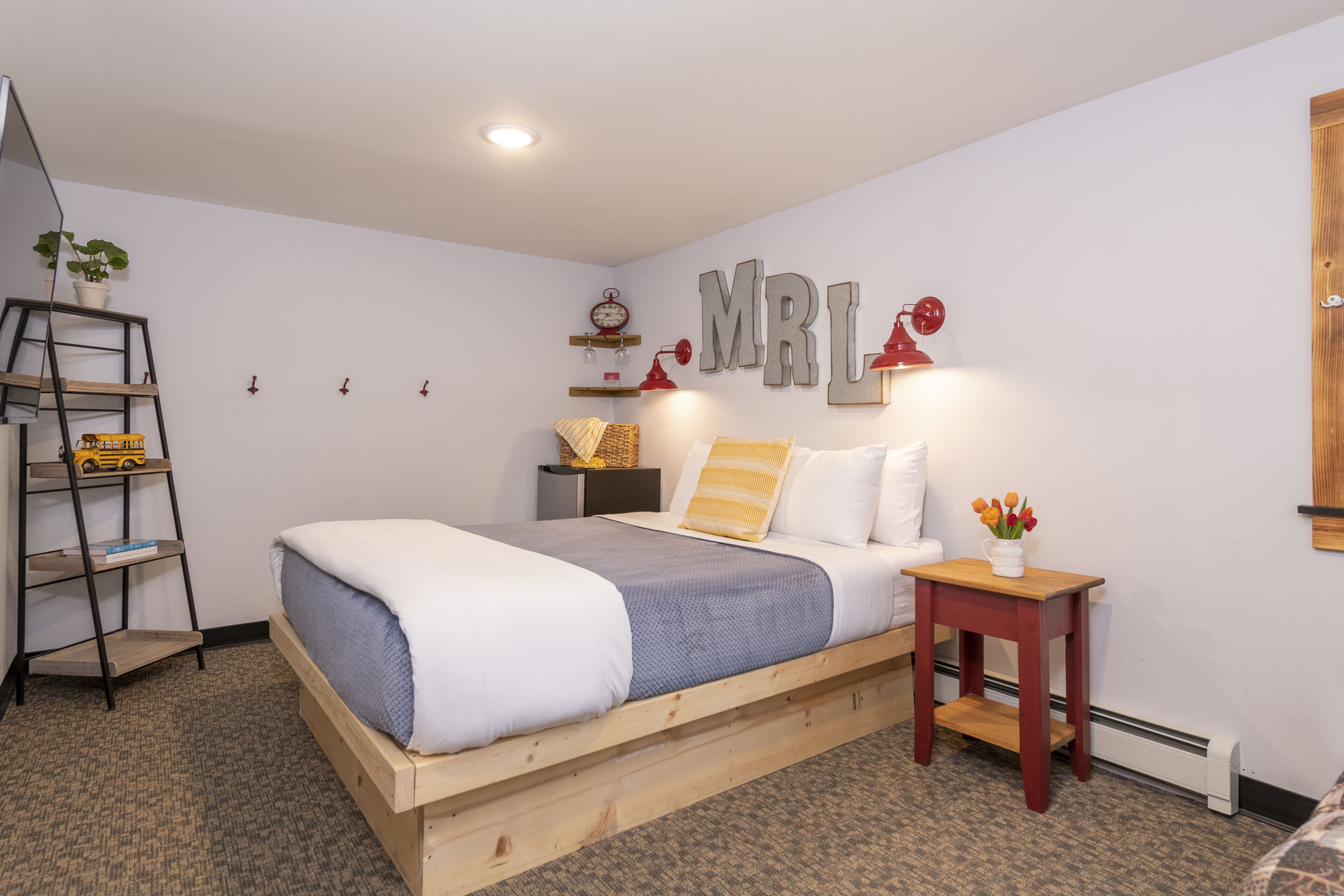 A bedroom with a wooden bed, a bedside table with a lamp, a shelf with decorative items, and a wall-mounted clock.