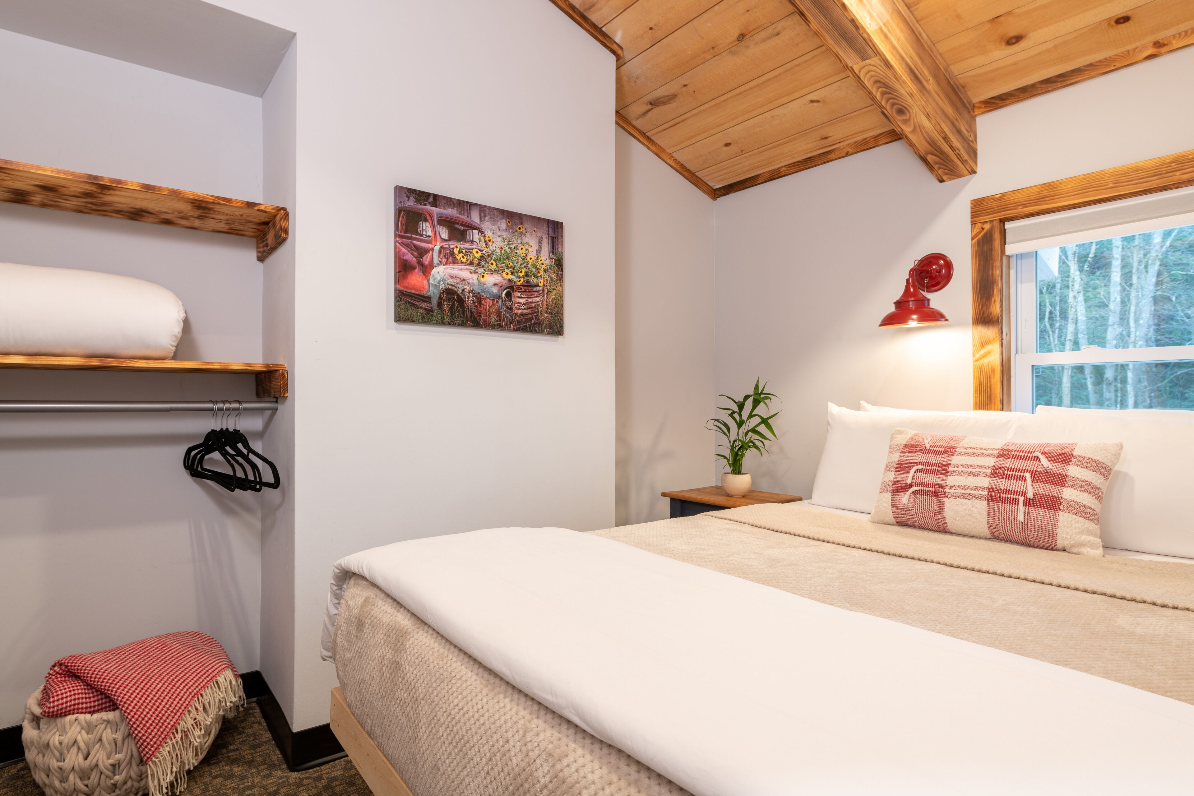 A bedroom with a wooden ceiling, a bed with a red lamp, a window with a view of trees, and a painting on the wall.