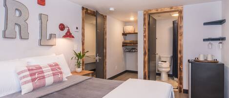 Una habitación de hotel moderna con una cama grande, una mesita de noche con lámpara, una planta pequeña y un refrigerador.