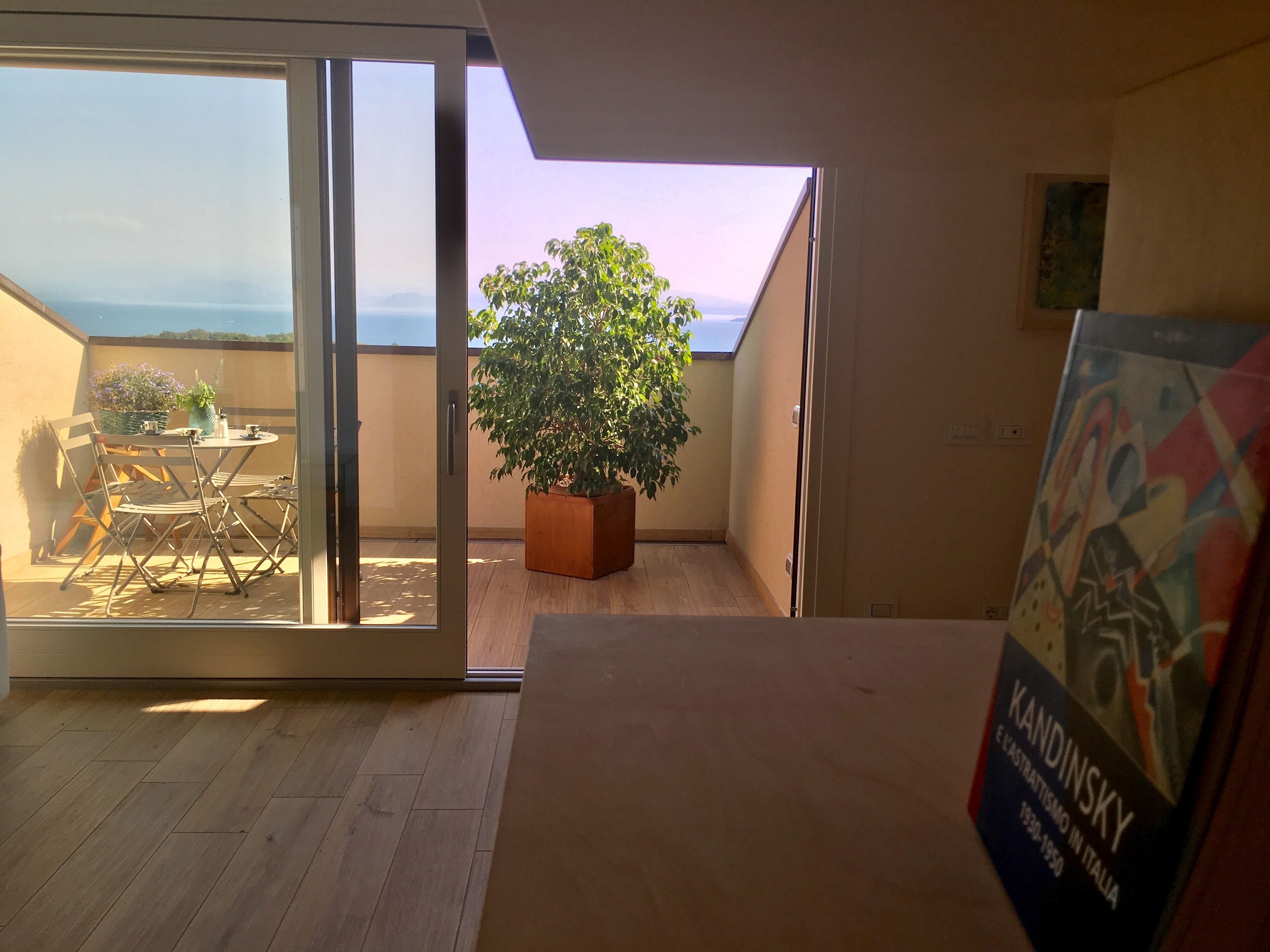 A room with a sliding glass door leading to an outdoor area with a table and chairs, a potted plant, and a view of the sea.
